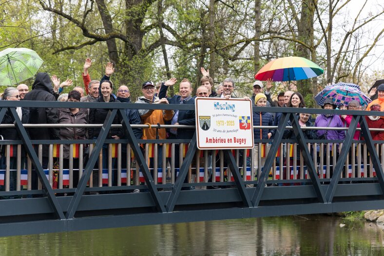 Viele Leute stehen mit dem Bürgermeister Florian A. Mayer auf einer Brücke mit dem Schild für die Partnerschaft mit Amberieu